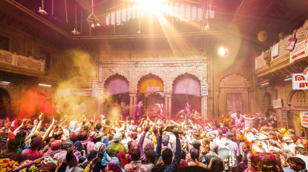 Multitud celebrando Holi en el templo, levantando los brazos bajo un rayo de sol mientras el aire se llena de polvos de colores vibrantes.