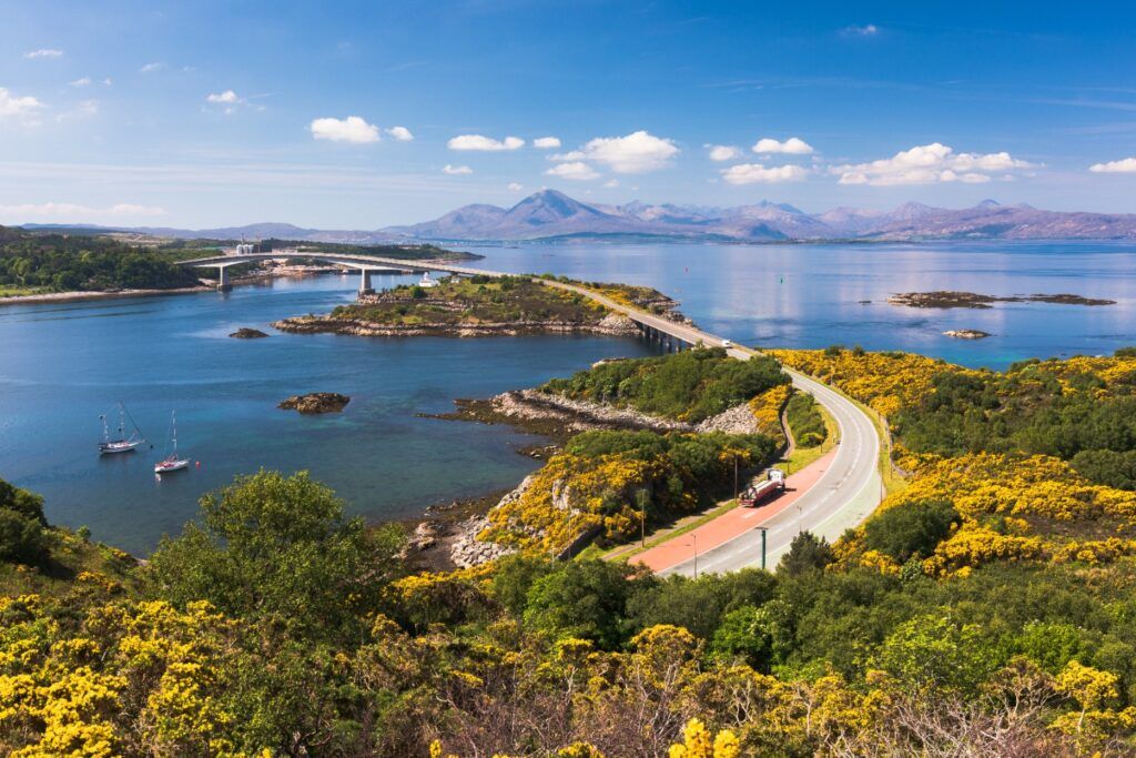 Puente de Skye (Skye Bridge) en día soleado, con carretera serpenteante, vegetación amarilla en flor y las montañas Cuillin al fondo.
