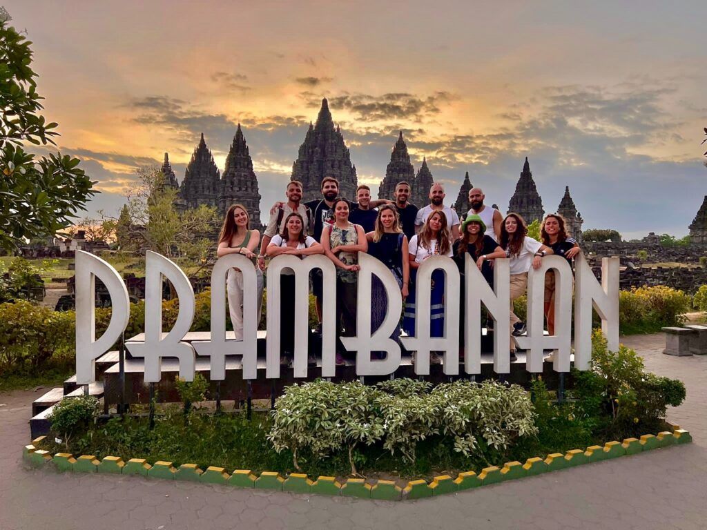 Grupo de viajeros WeRoad posando frente al cartel de Prambanan al atardecer.