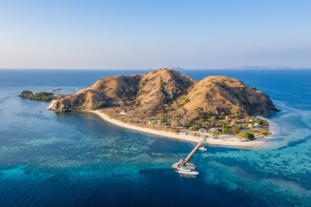 Toma aérea de una pequeña isla desértica de colinas marrones, con una playa de arena blanca, un pequeño pueblo costero y un muelle largo en aguas turquesas y azules.