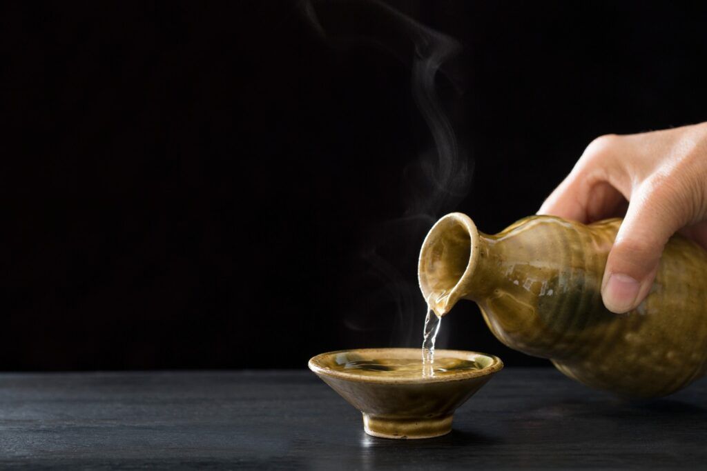Mano sirviendo sake caliente humeante (atsukan) en una taza tradicional japonesa.