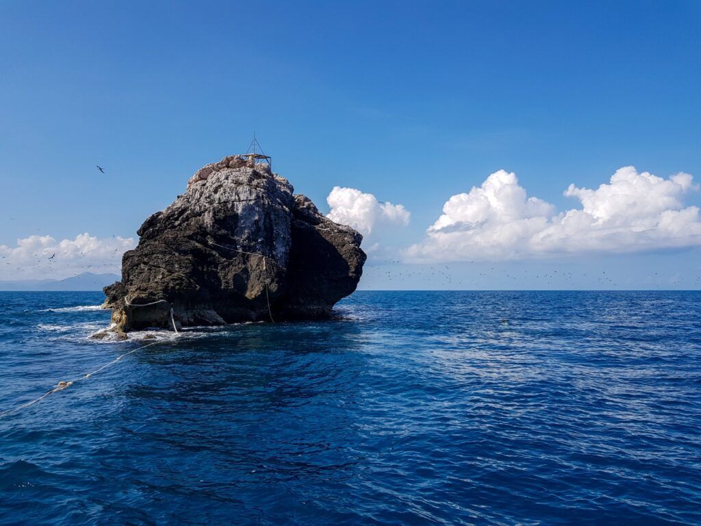 Roca solitaria emergiendo del mar, rodeada de aves marinas y aguas profundas, sitio de buceo Hin Daeng, Tailandia.