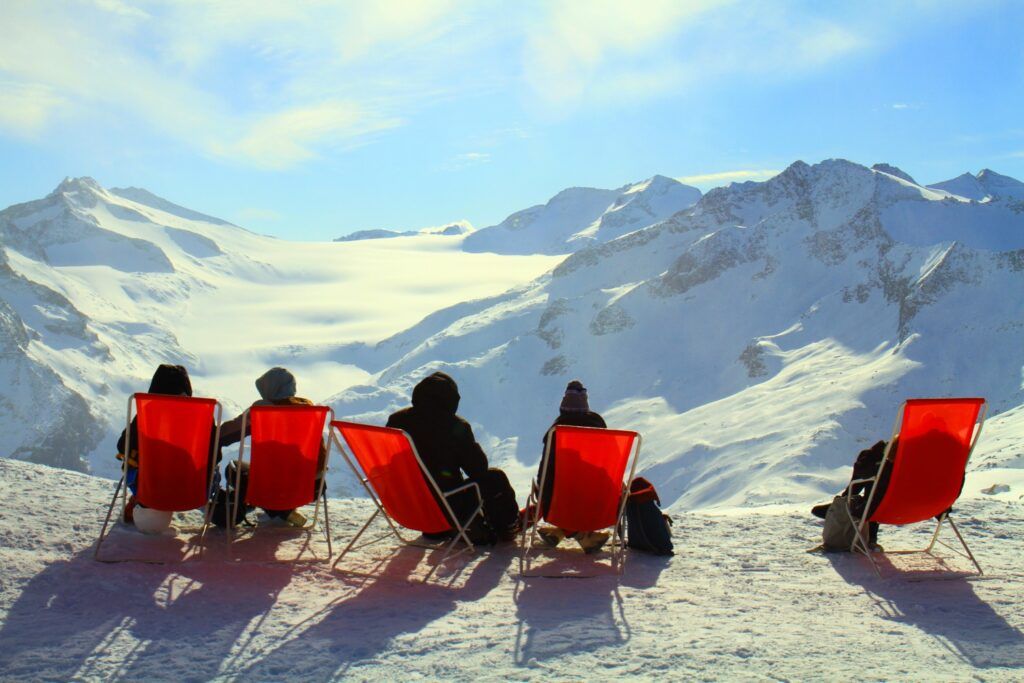 Momento après-ski: personas descansando en sillas de playa rojas en la nieve, admirando el valle y los picos nevados.