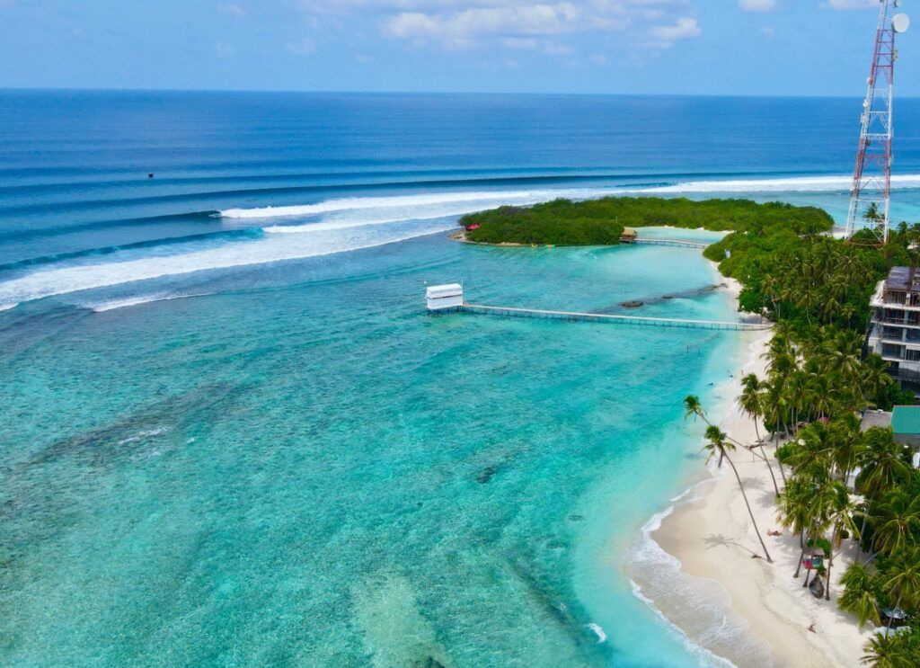 Vista panorámica desde el aire de una playa de arena blanca con palmeras inclinadas, aguas cristalinas y olas rompiendo a lo lejos en el océano.