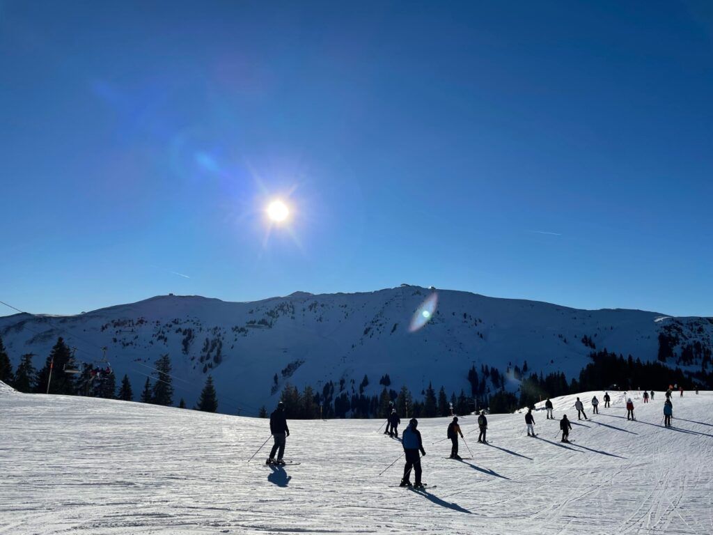 Amplia pista de esquí en Kitzbühel, Austria, con varios esquiadores bajo un sol alpino brillante y montañas al fondo.