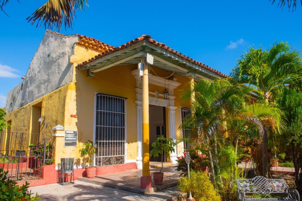 Casa colonial tradicional de color amarillo con portal de columnas, rodeada de vegetación tropical.