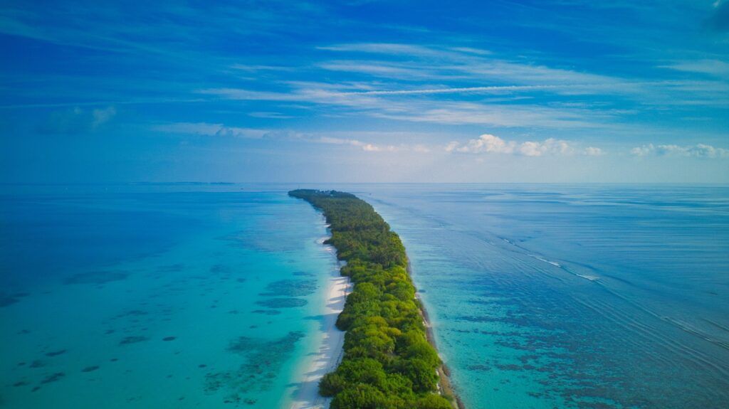 Vista aérea de una larga y estrecha lengua de tierra cubierta de vegetación tropical, rodeada por un arrecife de coral y aguas de color azul intenso.
