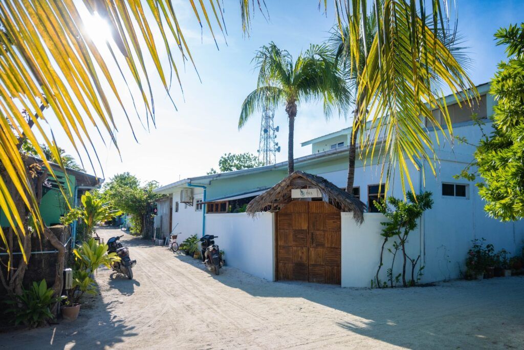 Una calle de arena blanca en una isla local de Maldivas con casas de colores, palmeras, motos aparcadas y una torre de comunicaciones al fondo bajo un sol brillante.