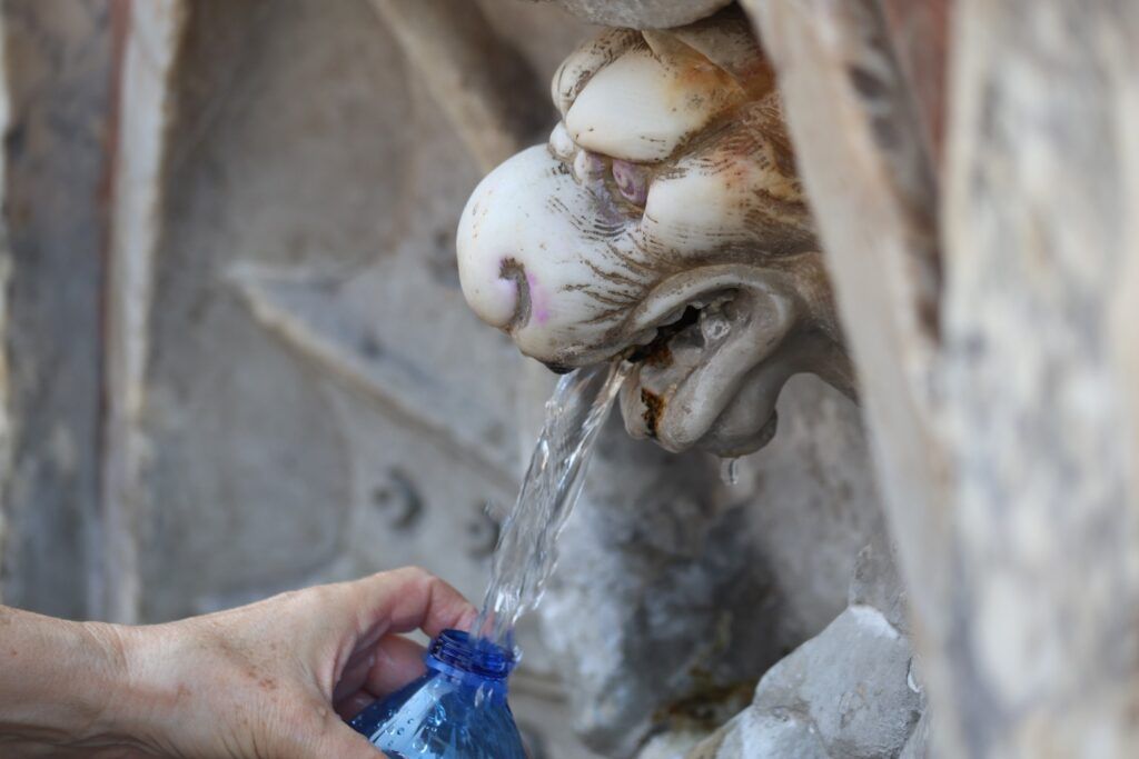 Primer plano de una mano llenando una botella de agua en una fuente histórica con forma de cabeza de león en Italia.