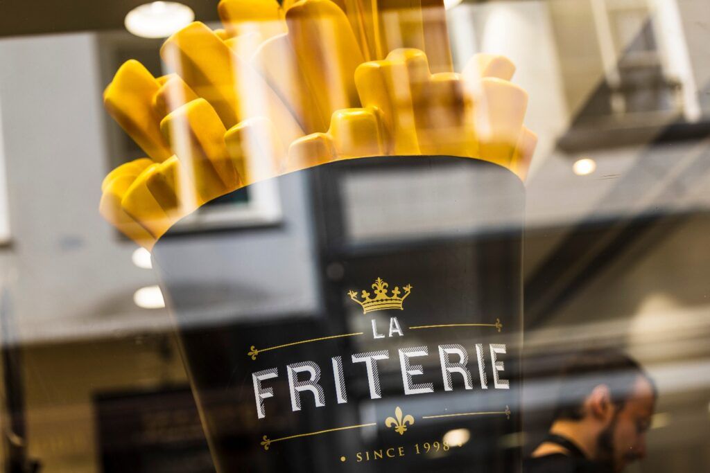Primer plano de un gran cartel o estatua de patatas fritas ('frites') en un envase negro con el logo 'La Friterie', visible a través de un cristal.