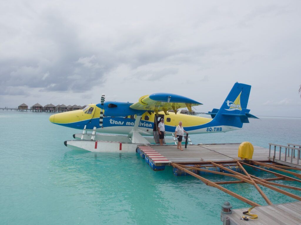 Un hidroavión azul y amarillo de Trans Maldivian Airways atracado en un muelle flotante de madera sobre aguas turquesas transparentes.