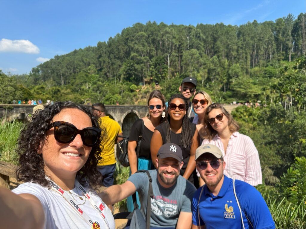 Un selfie de grupo de viajeros de WeRoad sonriendo frente al puente de los nueve arcos en Sri Lanka.