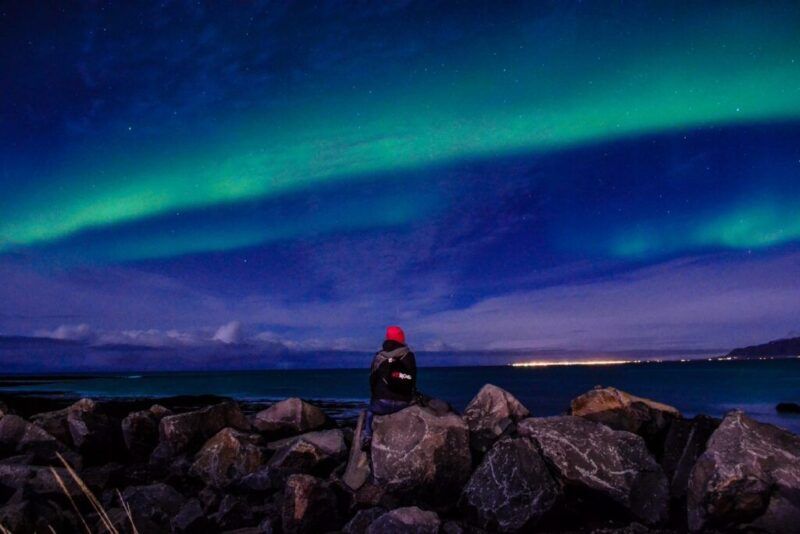 Una persona sentada en las rocas frente al mar bajo una banda de aurora boreal verde en Islandia.