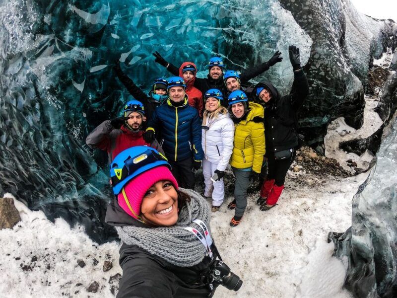 Un grupo de viajeros de WeRoad con cascos y equipo de montaña posa sonriente frente a una impresionante cueva de hielo azul en Islandia.