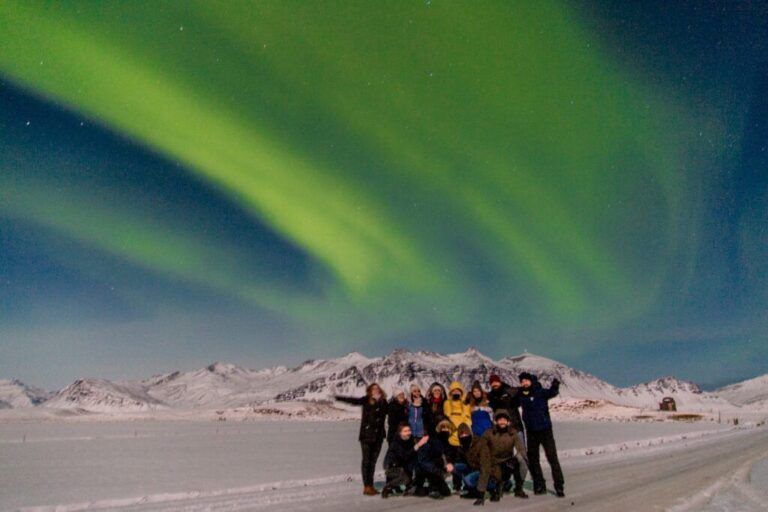 Un grupo de viajeros de WeRoad posa bajo una inmensa y brillante aurora boreal verde en un paisaje nevado de Islandia.