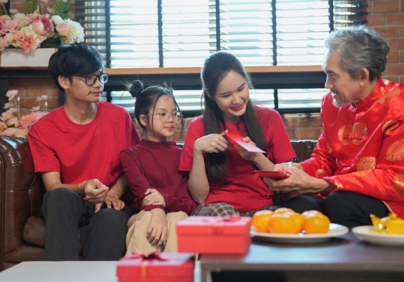 Una familia celebra el Año Nuevo chino en un salón decorado, donde un anciano ofrece sobres rojos tradicionales (hongbao) a unos jóvenes y a una niña.