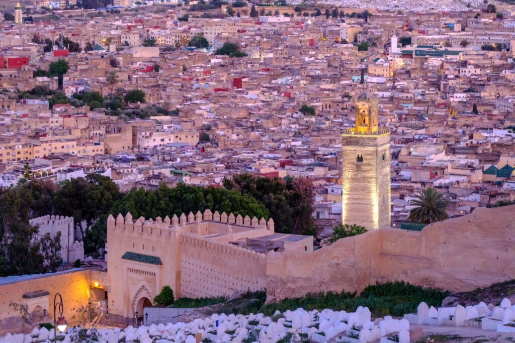 Vista panorámica al atardecer de una medina marroquí con casas de colores claros, una muralla antigua en primer plano y un minarete iluminado que destaca en el paisaje urbano.