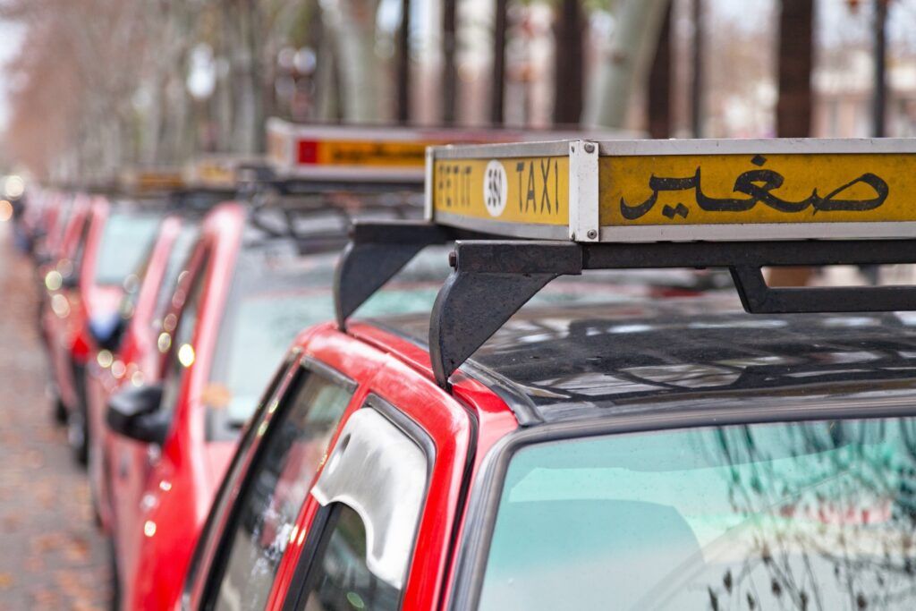 Fila de taxis rojos pequeños estacionados en una calle arbolada, con carteles amarillos en el techo que indican 'Petit Taxi' en francés y árabe.