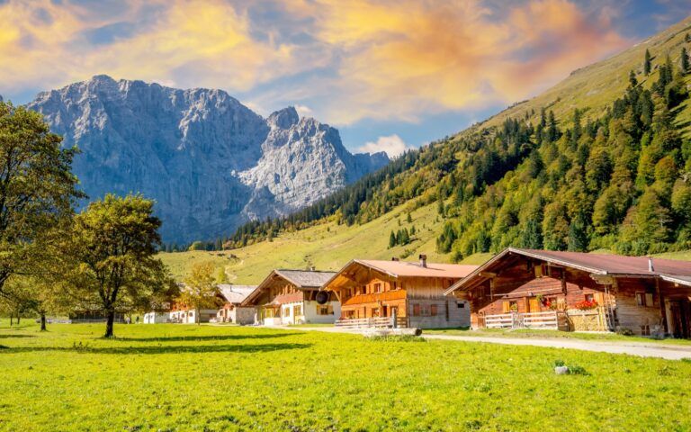 Un grupo de casas de madera tradicionales en un valle verde de los Alpes austriacos, con montañas escarpadas bajo un cielo cálido al atardecer.