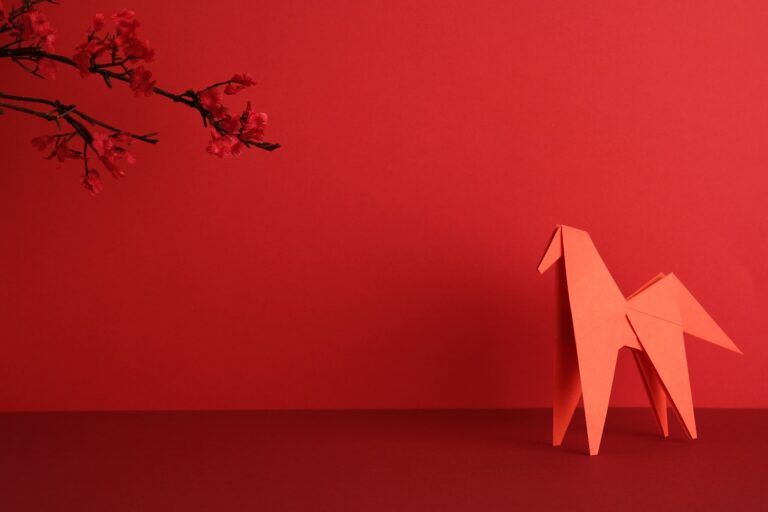 Un caballo rojo de origami colocado junto a una rama de cerezo en flor sobre un fondo rojo, celebrando el Año del Caballo para el Año Nuevo chino de 2026.