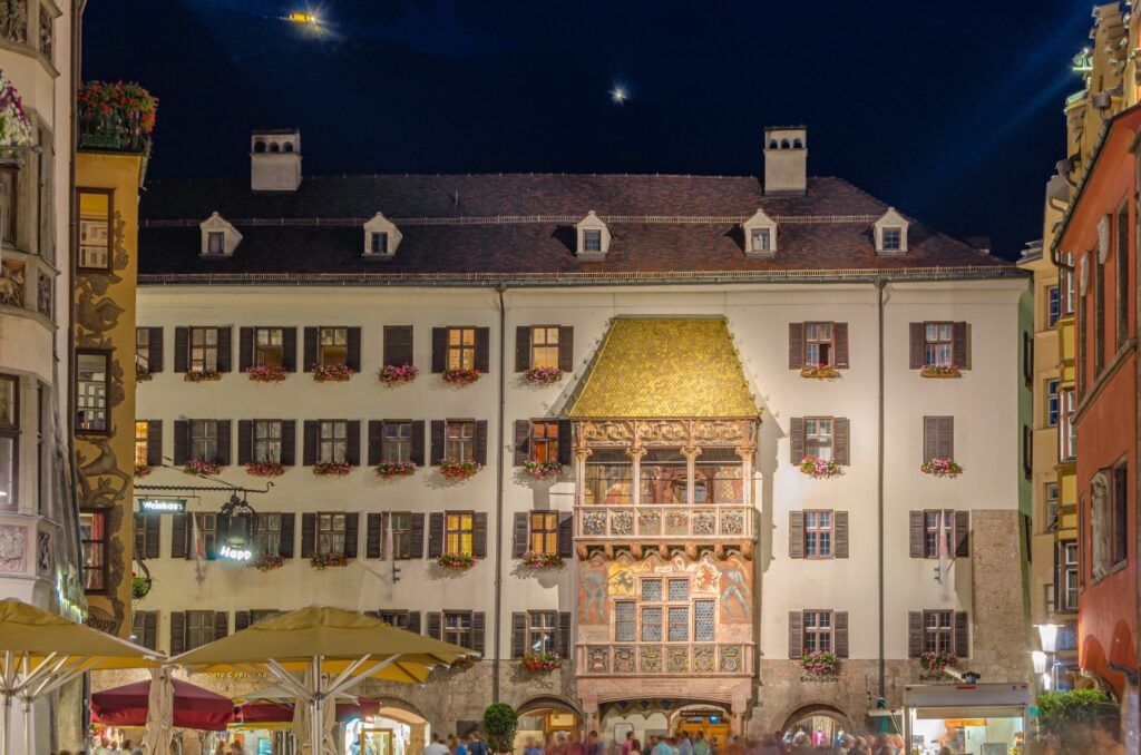 El emblemático Tejadillo de Oro de Innsbruck iluminado de noche, con su fachada decorada y ventanas llenas de flores en una plaza animada.