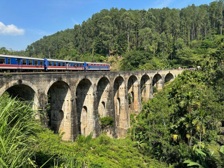Un tren azul y rojo cruza el histórico viaducto de piedra Nine Arch Bridge rodeado de selva en Sri Lanka.