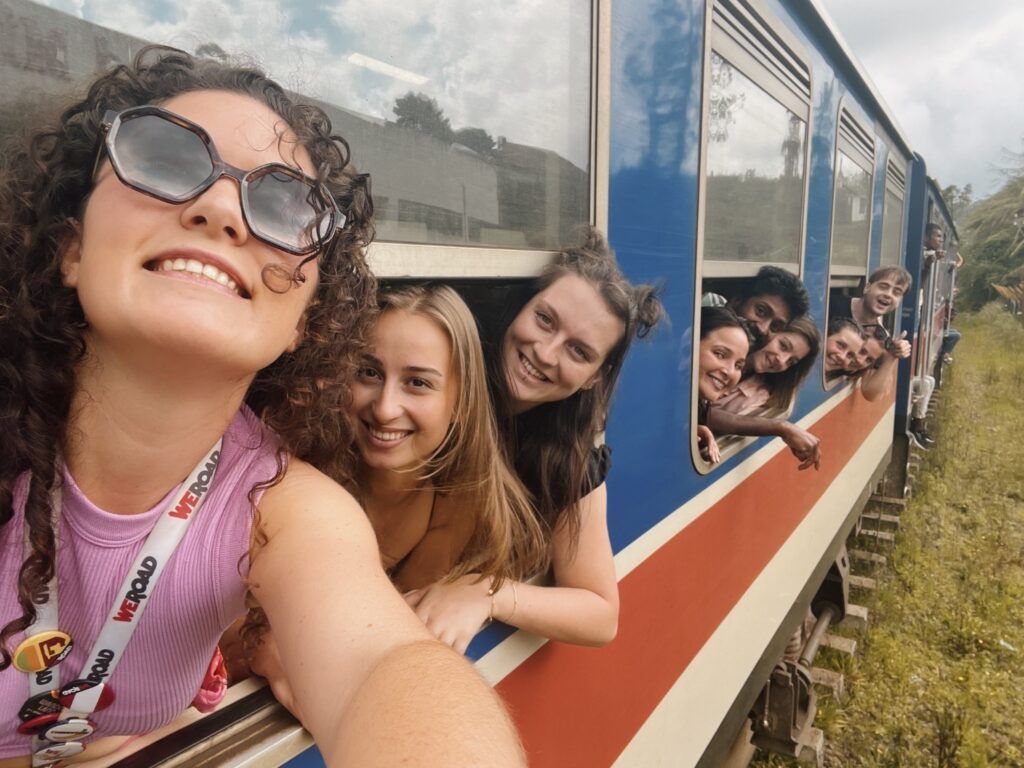 Un selfie de grupo de viajeros de WeRoad asomados por las ventanas de un tren azul en marcha en Sri Lanka.
