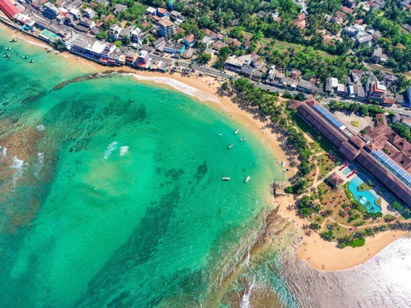 Vista aérea del mar turquesa y la playa de arena dorada de Hikkaduwa con palmeras y edificios.