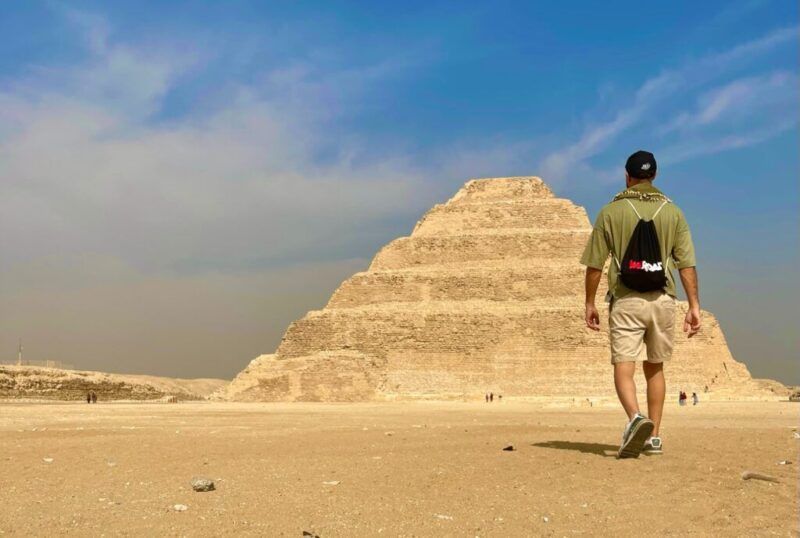 Un viajero con una mochila WeRoad camina hacia una pirámide escalonada en el desierto egipcio.