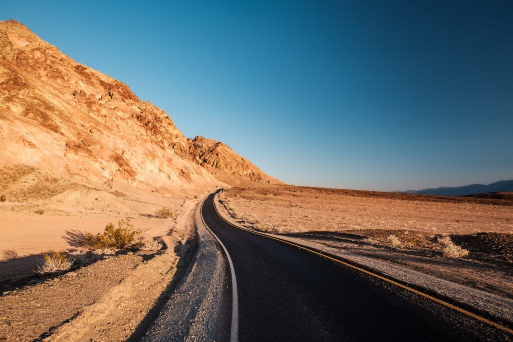 Una carretera asfaltada y sinuosa atraviesa el paisaje desértico de Artist's Drive en el Valle de la Muerte, rodeada de colinas rocosas bajo un cielo azul despejado.
