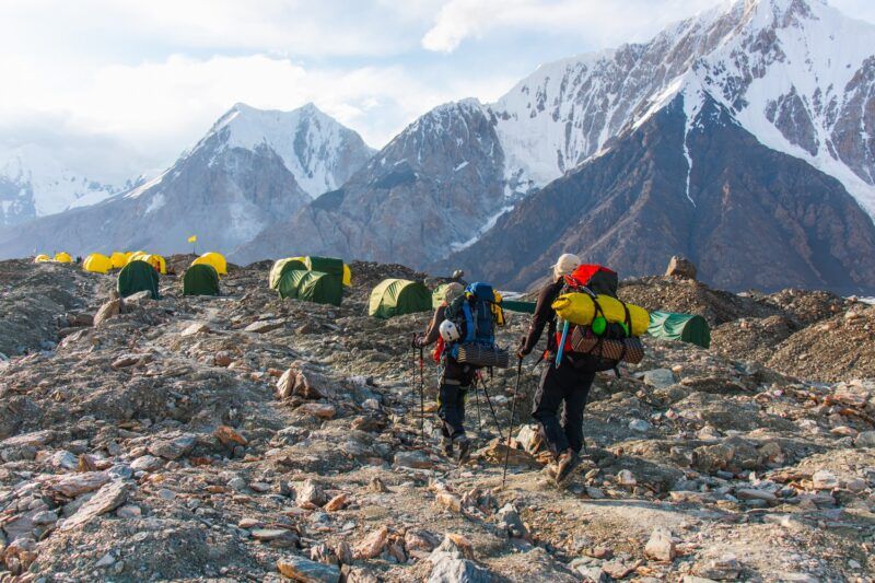 Dos senderistas caminan por un campamento de montaña a gran altitud, con tiendas amarillas y verdes frente a picos nevados.