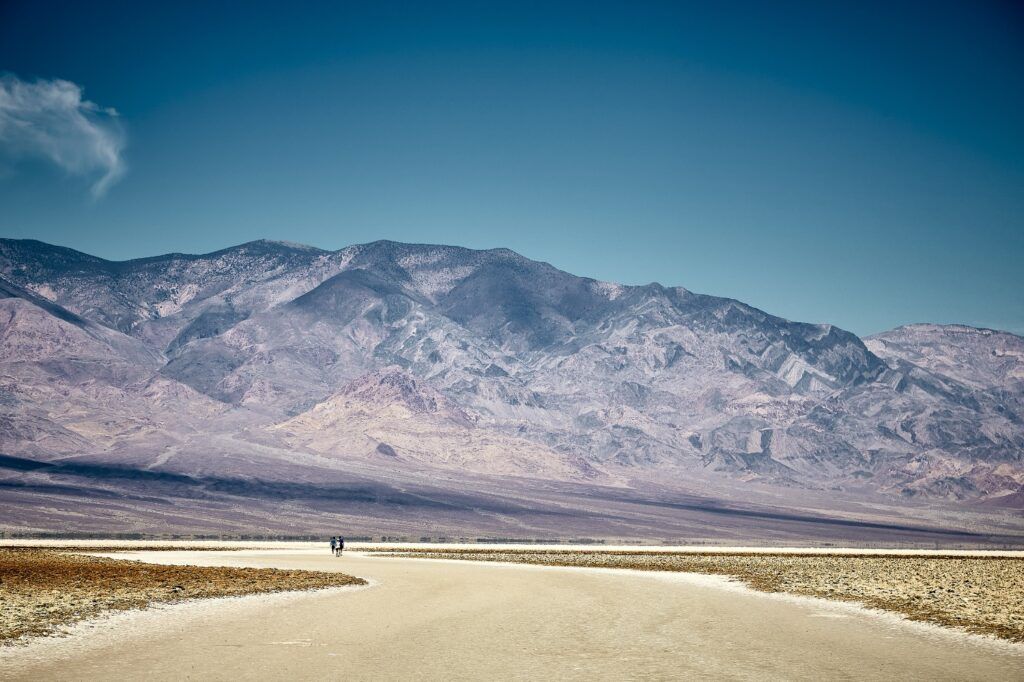 Dos personas caminan por el vasto y blanco salar de Badwater Basin, rodeadas de montañas desérticas bajo un cielo azul profundo en el Valle de la Muerte.