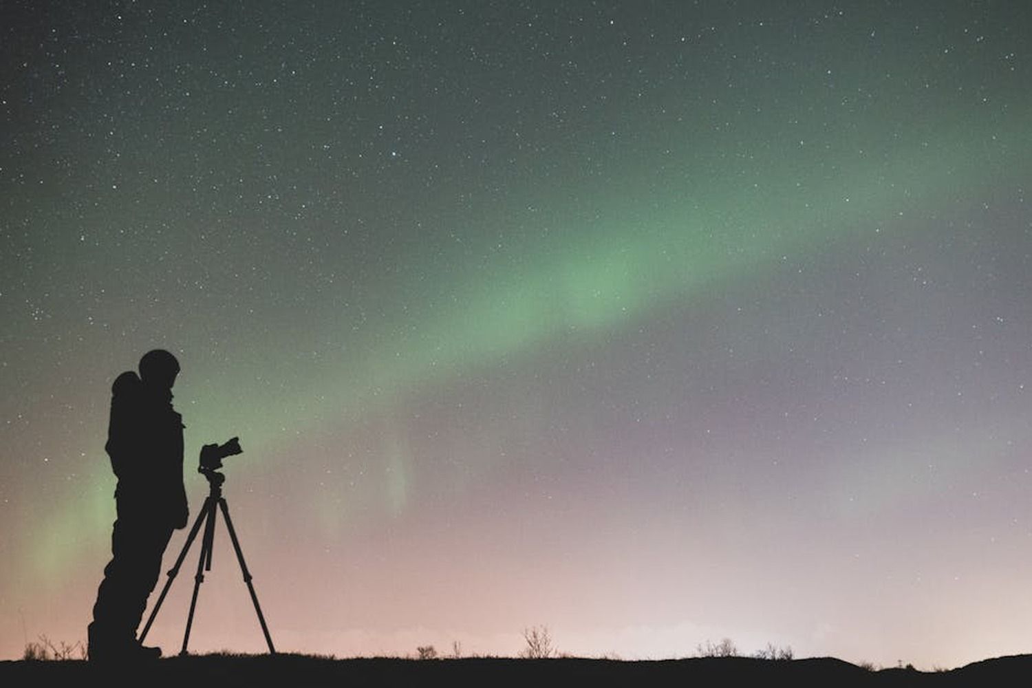 Silueta de un fotografo con tripode capturando la aurora boreal verde bajo un cielo estrellado nocturno