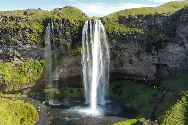 Cascada de Seljalandsfoss cayendo desde un acantilado cubierto de vegetación verde en Islandia, con turistas visibles al pie