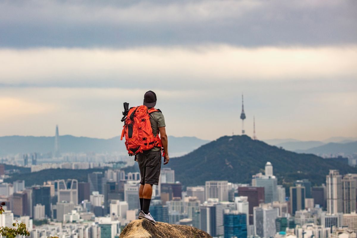 Hombre con mochila roja de pie sobre una roca observando la ciudad desde la cima