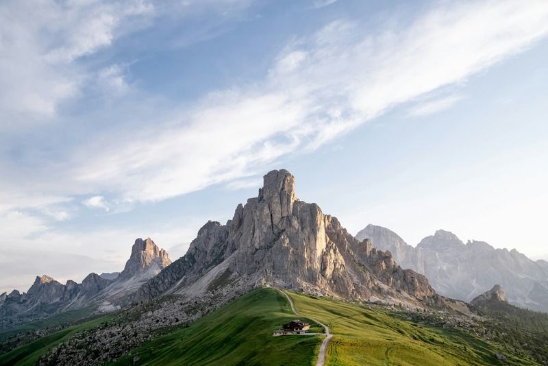 Refugio de montaña junto a un sendero serpenteante en una colina verde, con picos rocosos de los Dolomitas al fondo
