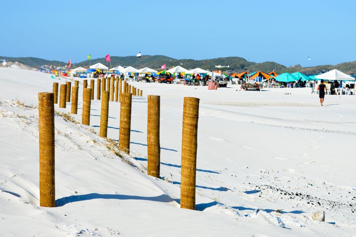 Playa de arena blanca con postes de madera, carpas de colores y colinas verdes al fondo