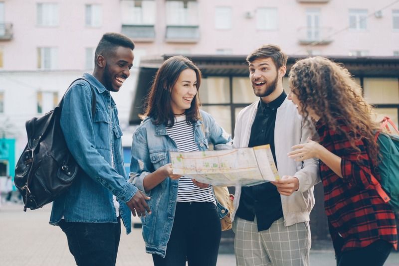 Cuatro amigos riendo mientras consultan un mapa de papel en una calle urbana con edificios de colores pastel