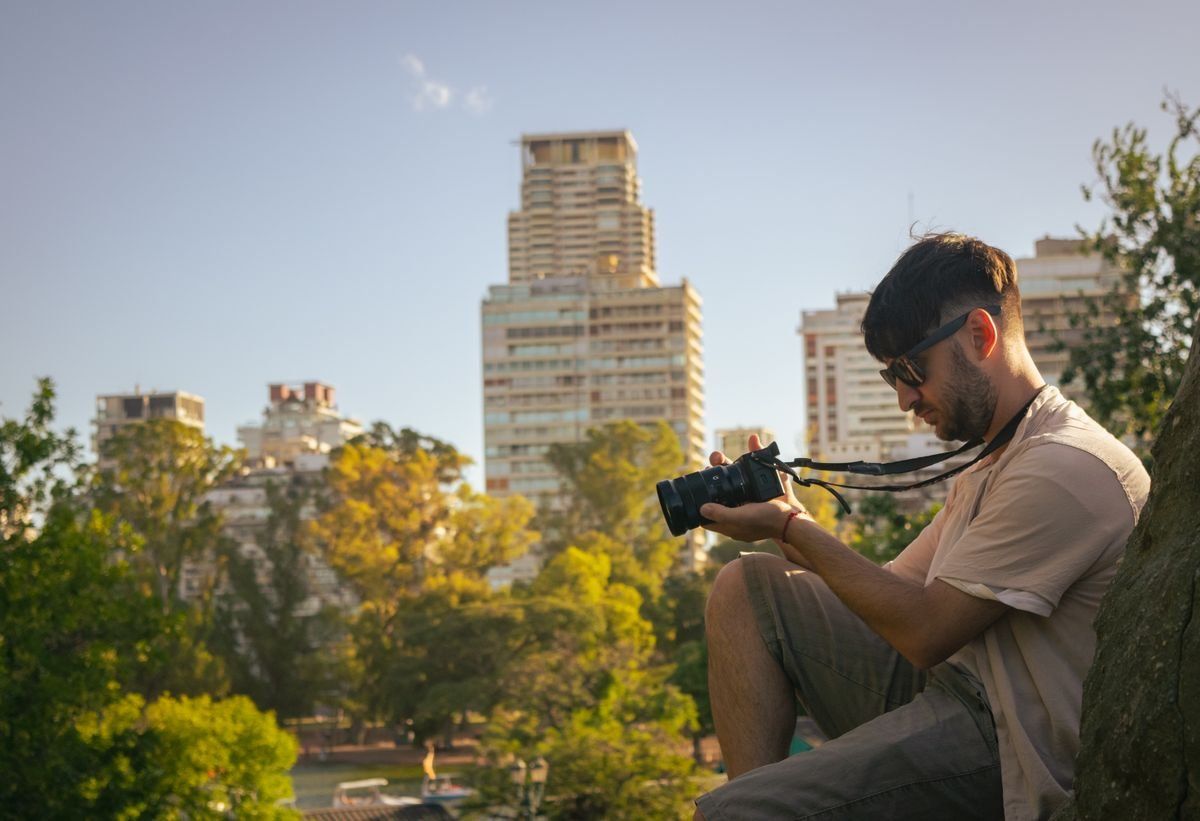 Fotógrafo sentado en un parque mirando su cámara con edificios al fondo