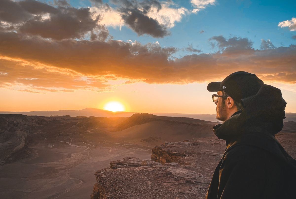 Hombre con gorra contemplando el atardecer sobre un paisaje desértico desde una roca