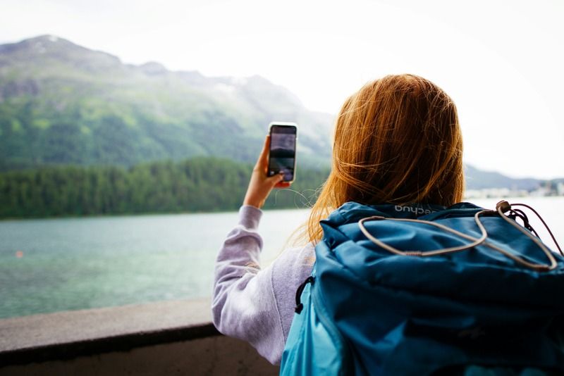 Mochilera fotografiando con su móvil un lago alpino turquesa rodeado de montañas verdes