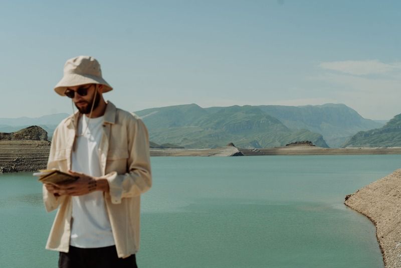 Un viajero con sombrero de pescador y gafas de sol leyendo un cuaderno junto a un embalse turquesa rodeado de montañas verdes