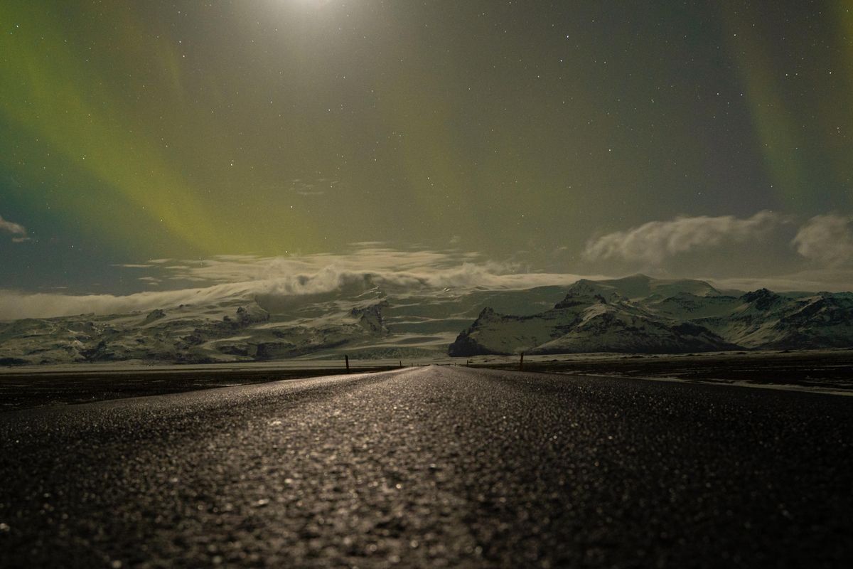 Carretera oscura bajo aurora boreal con montañas nevadas en la distancia