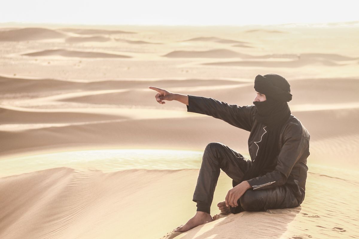 Hombre con ropa tradicional sentado en una duna señalando el horizonte