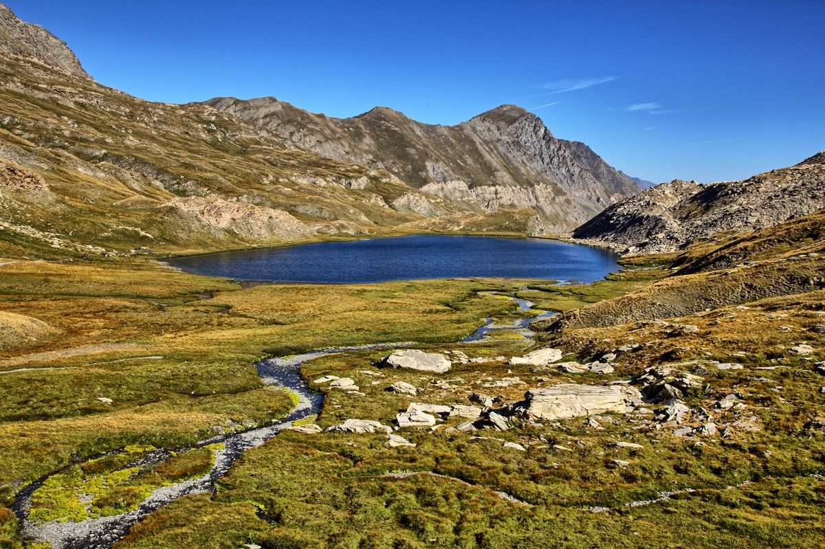 Lago de montaña rodeado de rocas, praderas y un arroyo serpenteante