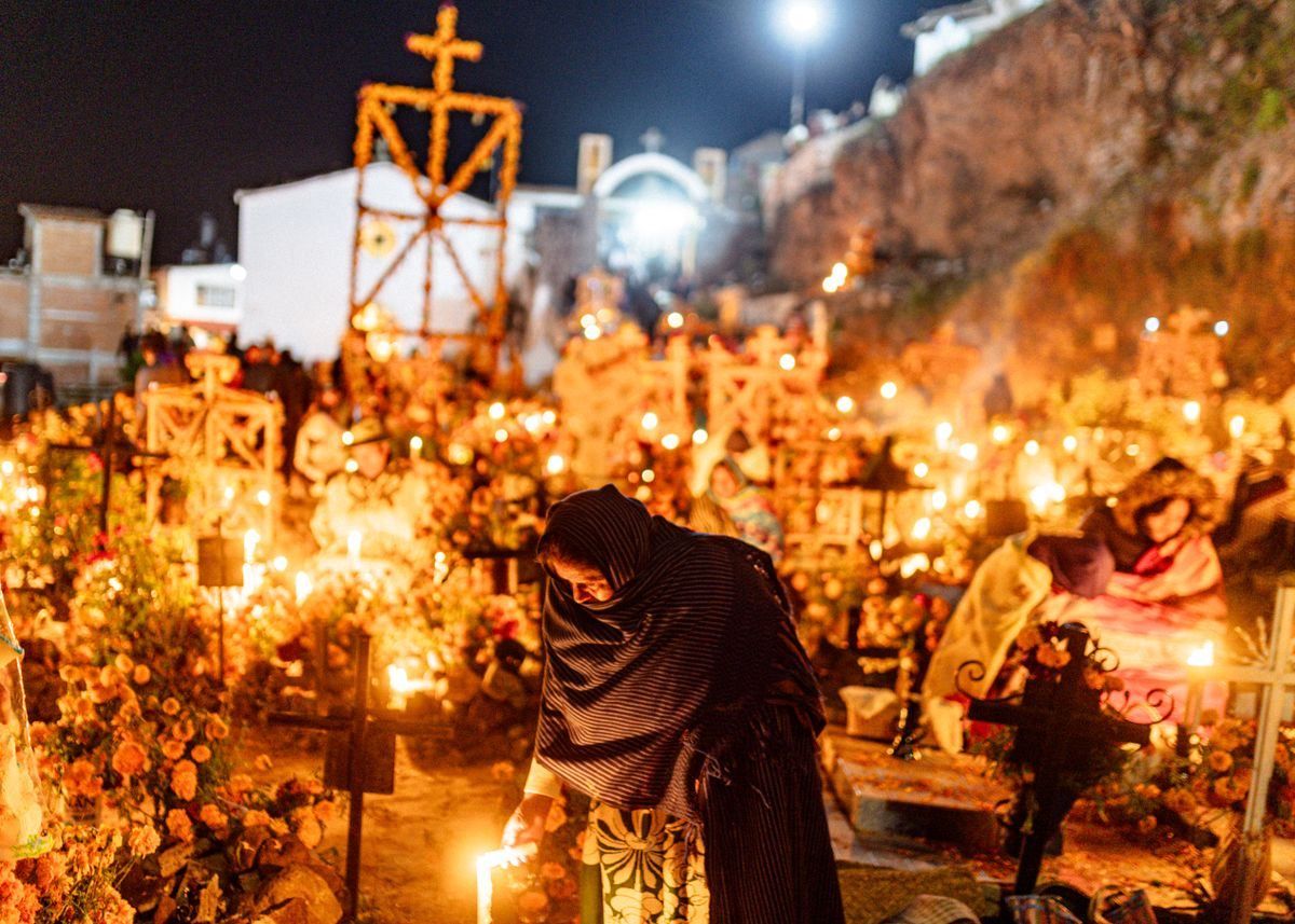 Mujer encendiendo una vela entre tumbas cubiertas de flores y luces en un cementerio nocturno