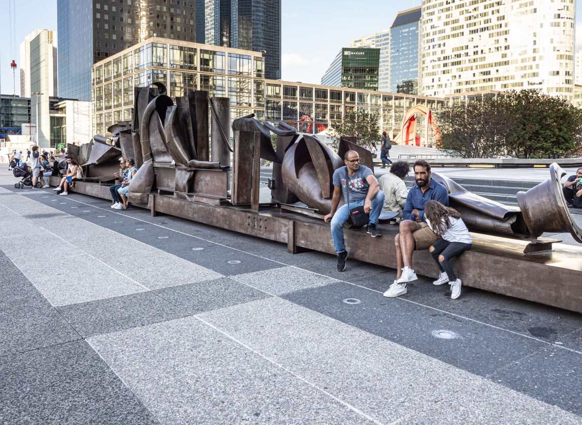 Personas sentadas junto a una escultura moderna en La Défense con rascacielos al fondo