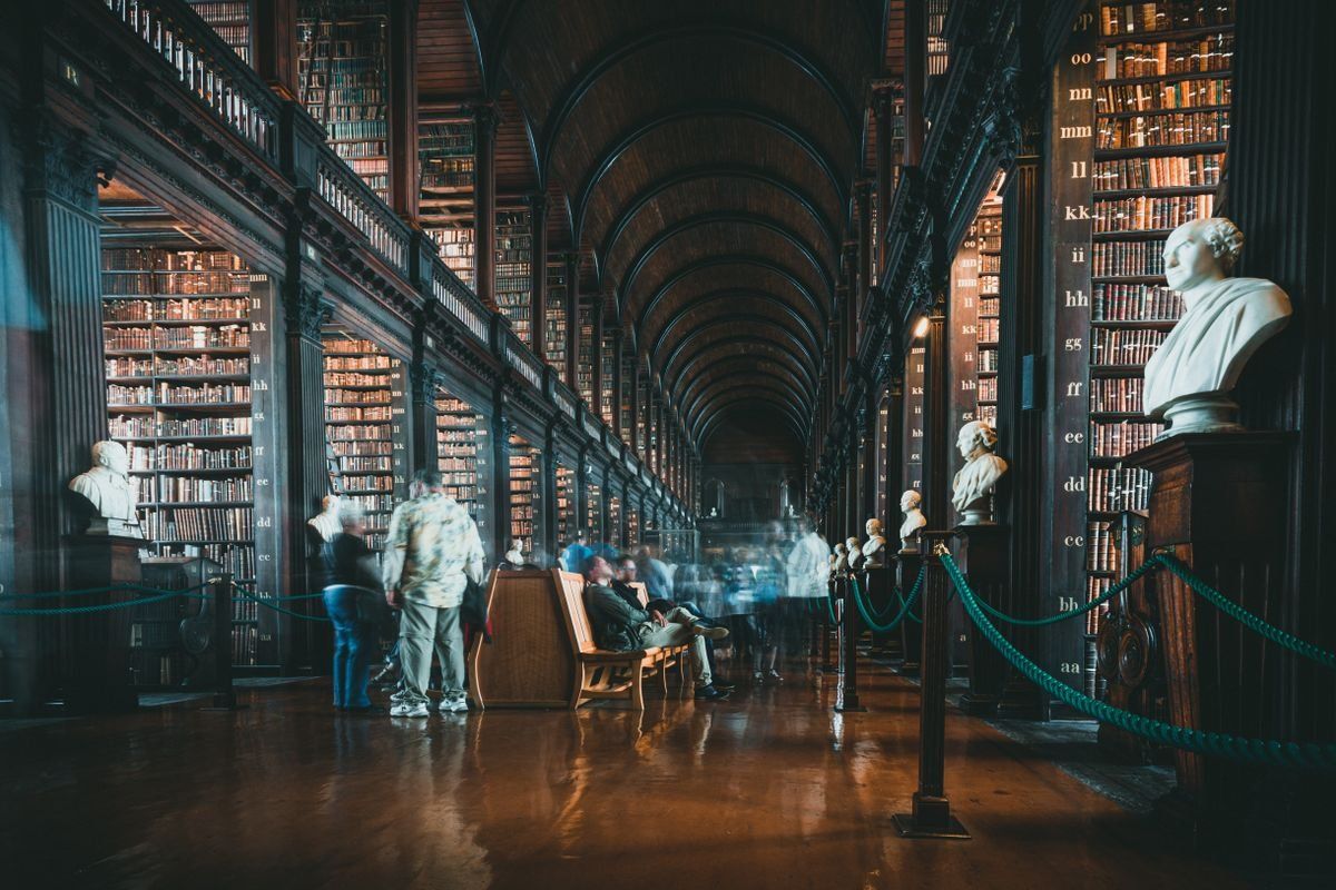 Larga sala de biblioteca con estanterías altas, bustos de mármol y personas desenfocadas